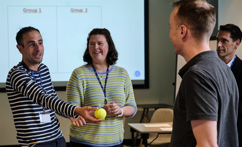 Workshop participants engaging in interactive exercise with tennis ball, wearing striped shirts and lanyards, presentation screen showing 'Group 1 Group 2' visible in background