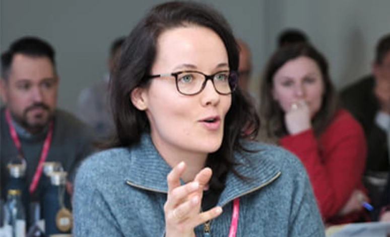 Person in grey sweater actively speaking during a business meeting, wearing glasses, with a lanyard badge and reusable water bottle on the table