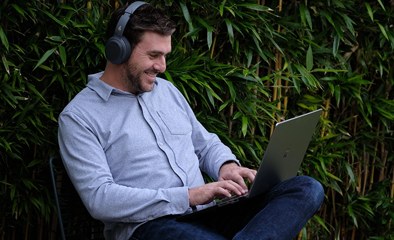 Smiling professional working outdoors on laptop while wearing headphones, seated against bamboo background in casual grey shirt and jeans