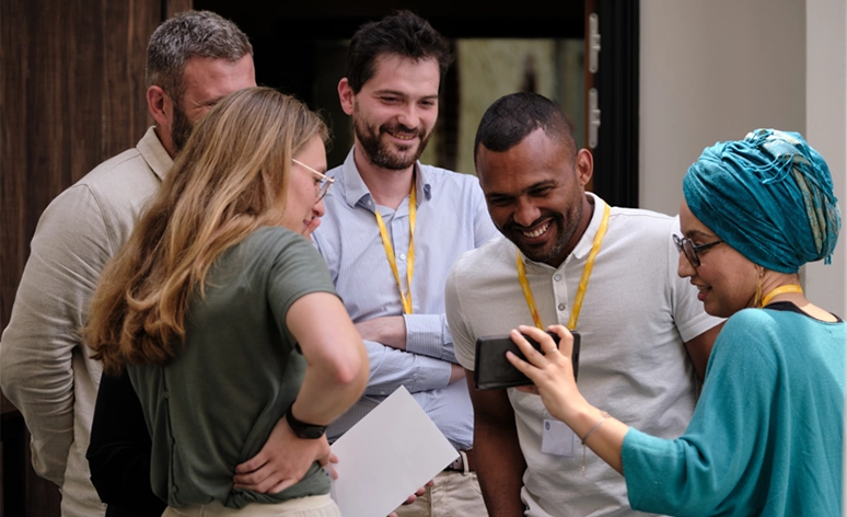 Group of diverse professionals wearing lanyards sharing a lighthearted moment while looking at mobile device, dressed in casual business attire against wooden doorway backdrop