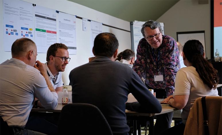 Workshop session with participants seated around table, person in floral shirt standing and engaging with group, white board with documents visible in background