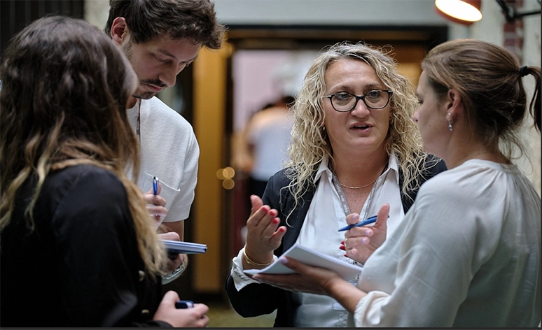 Professional group discussion in office hallway with four people taking notes, focused on person with curly blonde hair and glasses in business attire gesturing while speaking