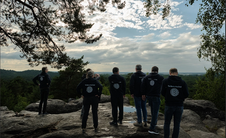 Group of six people wearing matching black hoodies with logos standing on rocky outcrop overlooking scenic landscape with dramatic cloudy sky