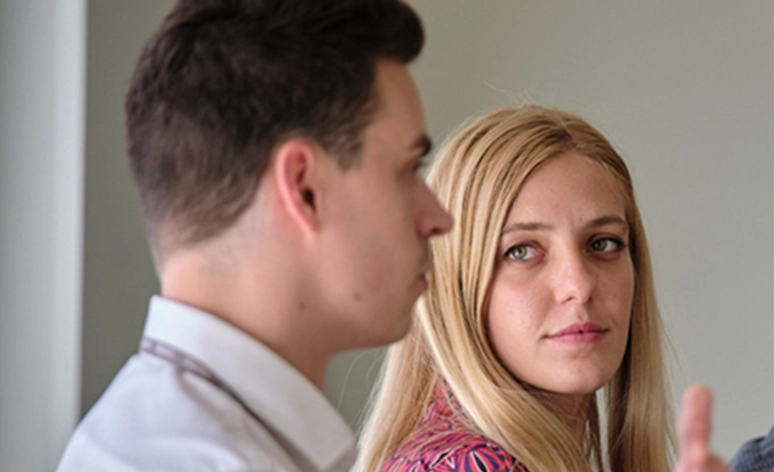 Professional conversation between two colleagues in close profile view, one person looking directly at camera while other person gestures mid-discussion