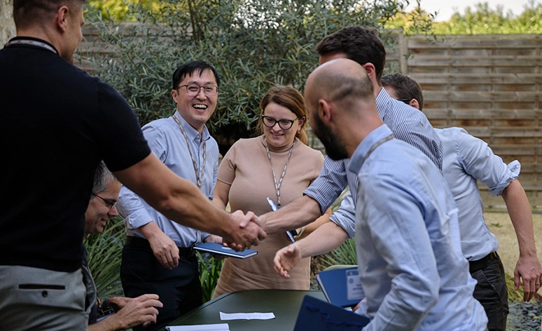 Group of professionals celebrating with a handshake during outdoor meeting, team members smiling and wearing lanyards, gathered around workspace in garden setting